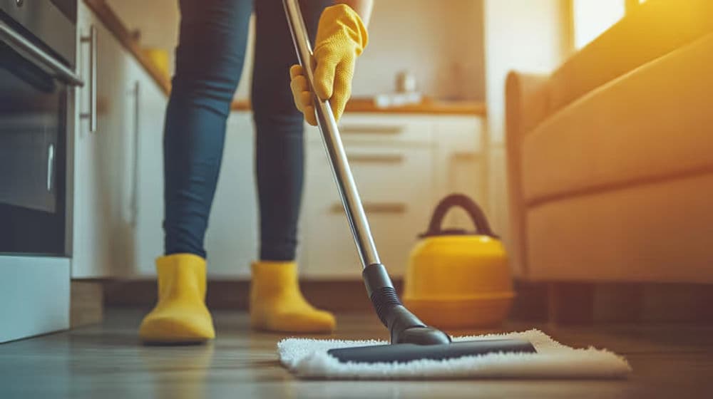 Person in yellow gloves mopping a kitchen floor with a vacuum cleaner in the background.